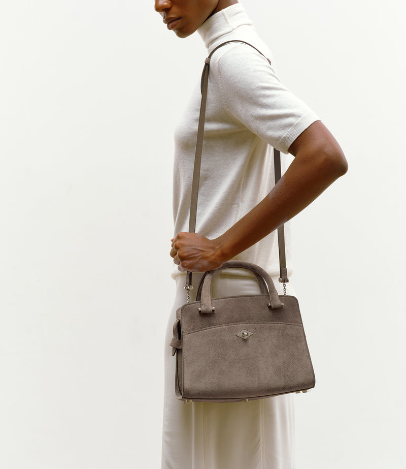 A model wearing a white turtleneck sweater and white trousers is shown from the side, carrying a taupe-brown suede crossbody handbag with a top handle and shoulder strap against a plain off-white background.